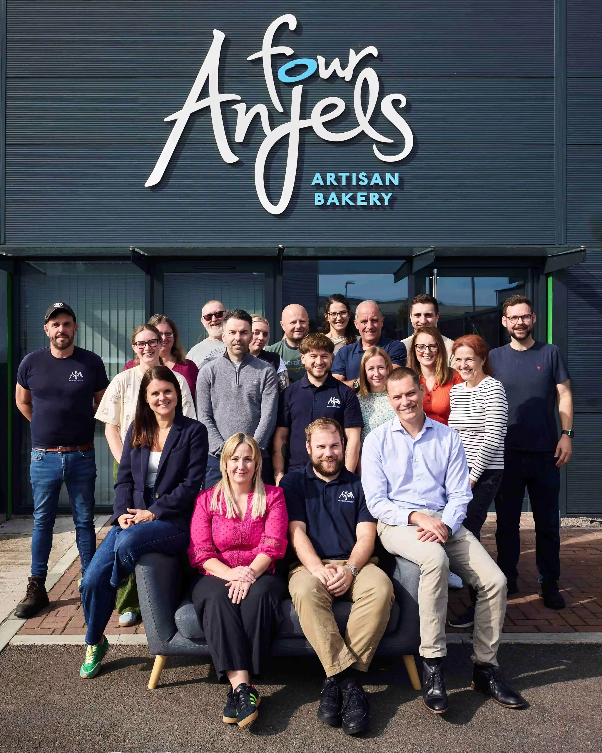 A group of people stand and sit smiling at the camera. They are in front of a building with a sign that reads Four Anjels
